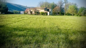 A rural landscape with a large grassy field, a traditional stone house, and a mountain in the background.