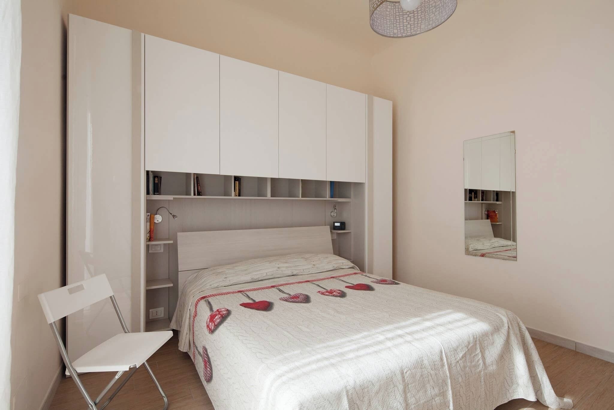 Ein Schlafzimmer mit Bett, Sessel, Kleiderschrank, Regal und Spiegel.