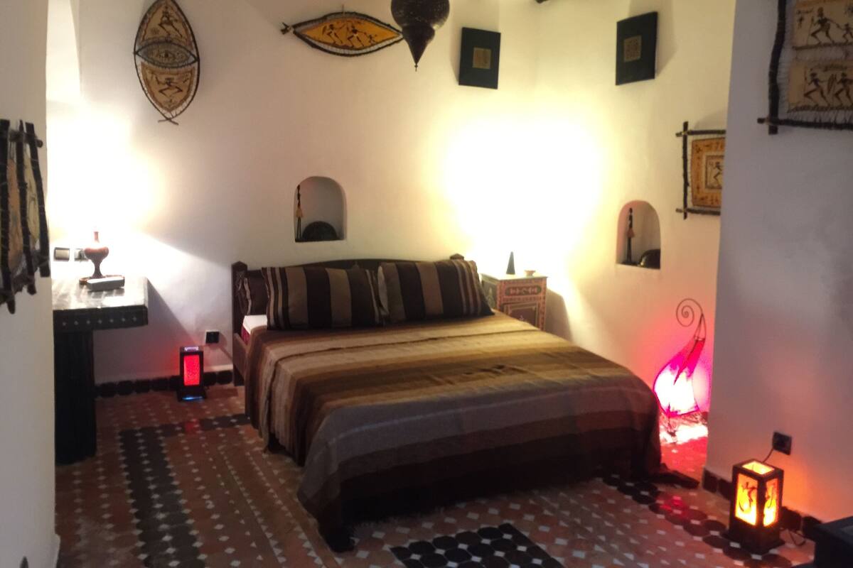 A bedroom with a bed, a nightstand, a wall-mounted shelf, and a patterned floor.