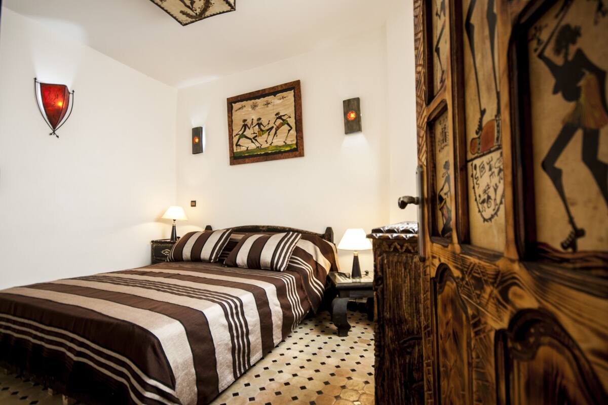 A hotel room with a bed, a nightstand, a lamp, and a wooden door with artwork.