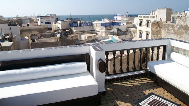 A rooftop terrace with white sofas and a patterned floor, overlooking a coastal cityscape with buildings and the sea. - Riad Bab Essaouira (Essaouira)