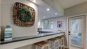 Een keuken met een granieten aanrecht, witte barkrukken, een geweven mand van hout met groen, en een schilderij van bloemen.