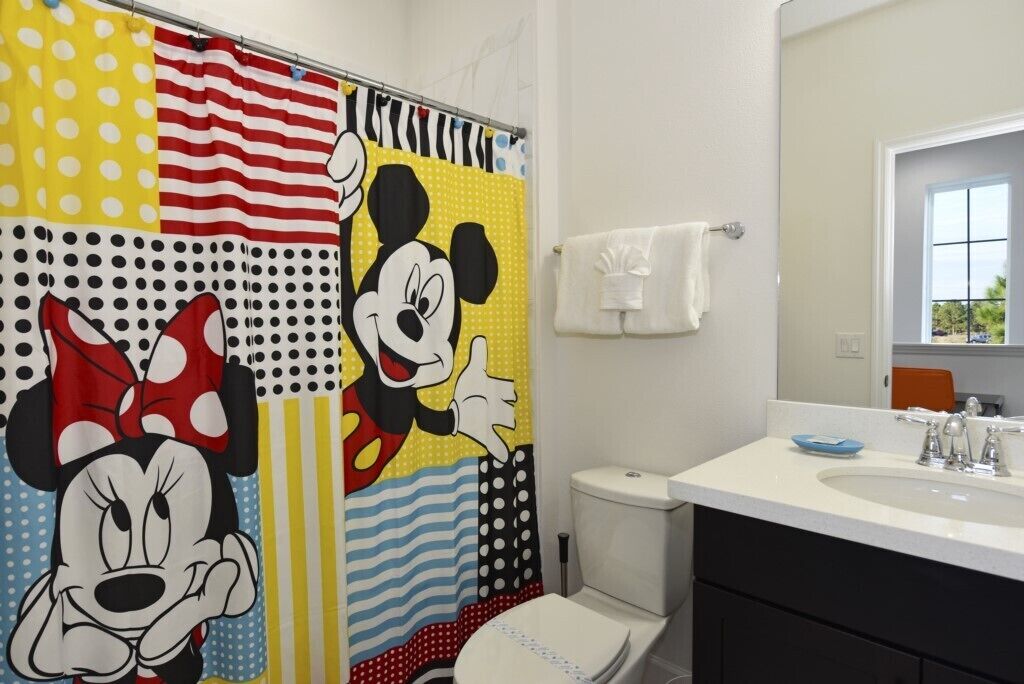Ein Badezimmer mit einer Mickey Mouse Duschtuchvorhang, einer weißen Toilette, einem Waschbecken mit Spiegel und einem Handtuchhalter.