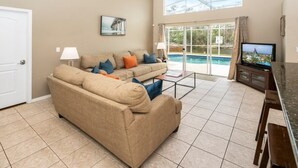 A living room with a beige sofa, a coffee table, a television, and a view of a swimming pool through large glass doors. - Wr001or 4 Bedroom Home by RedAwning (Davenport)