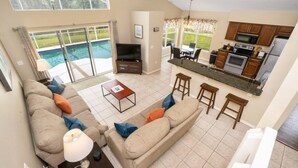 A spacious living area with a large sofa, a coffee table, and a TV. There is a kitchen with a dining area and a view of a swimming pool. - Wr001or 4 Bedroom Home by RedAwning (Davenport)