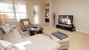 A living room with a beige sofa, a glass coffee table, a television, and a door leading to another room. - Vista Cay Gem 3 Bedroom Condo by RedAwning (Orlando)
