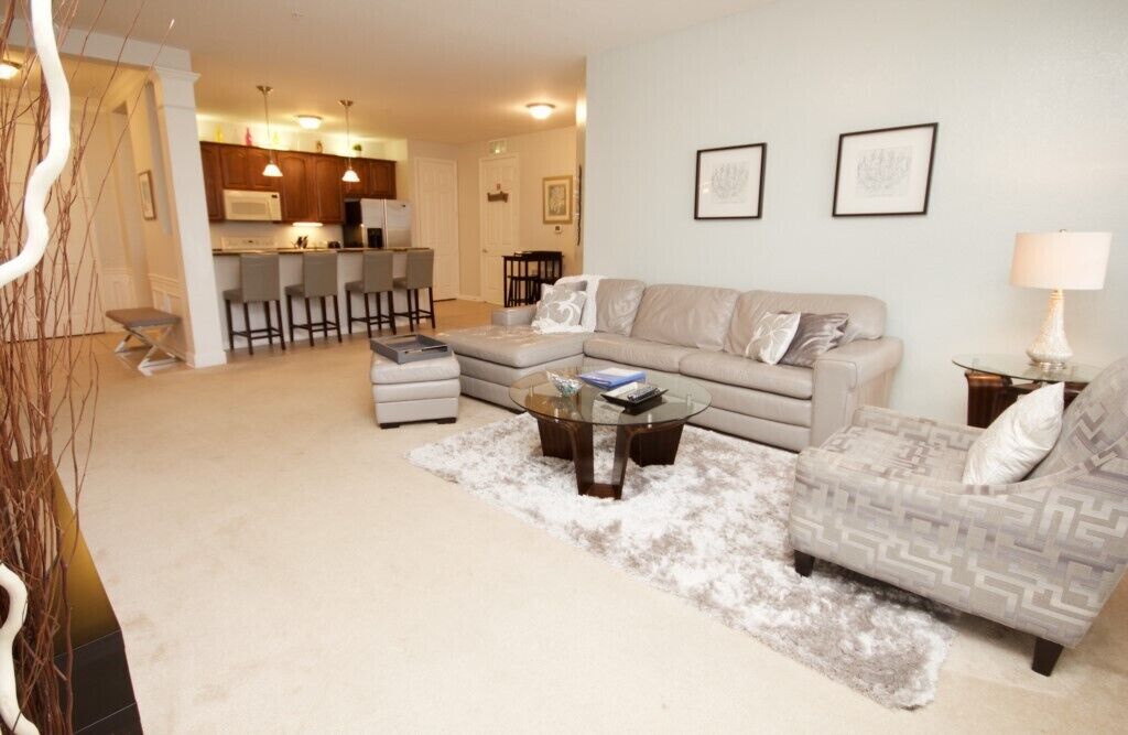 A living room with a sofa, ottoman, and coffee table, a kitchen in the background, and a dining area with bar stools.