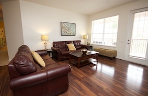 A living room with a brown leather sofa, a wooden coffee table, and a bench. There are two lamps on side tables, a painting on the wall, and a door with a window.