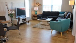 A modern living room with a flat-screen TV, a sofa, a coffee table, and a dining area with chairs.