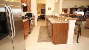A kitchen with wooden cabinets, a stainless steel refrigerator, a granite countertop island, and a dining area with wicker chairs.