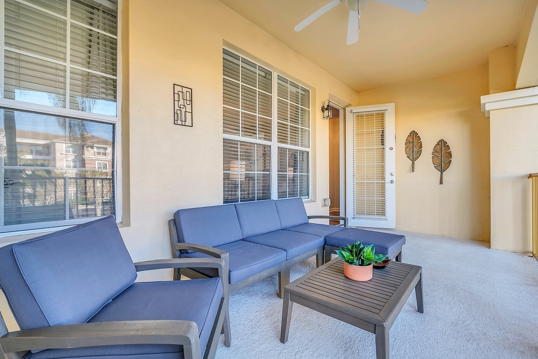 A sunlit patio with a blue sofa, a wooden table with a plant, and decorative leaf wall art.