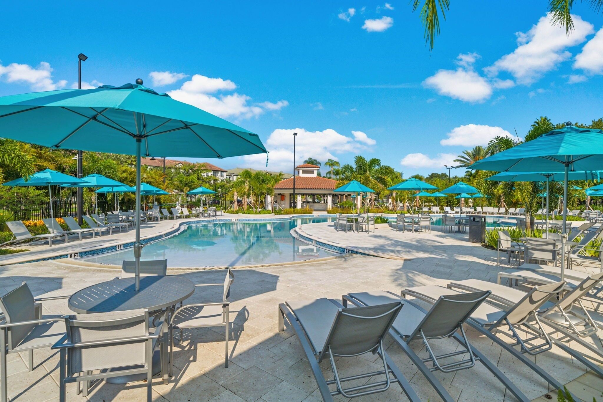 A pool area with lounge chairs and umbrellas, surrounded by palm trees and a clear blue sky.