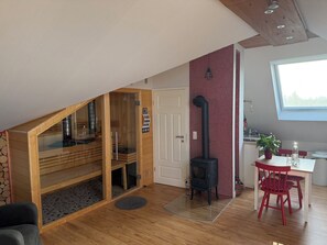 A cozy loft bedroom with a wooden sloped ceiling, a glass-walled shower enclosure, a black wood-burning stove, and a dining area with a red chair and table. - Wellnessloft mit Privatsauna und Garten (Ortenberg)