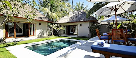 A pool area with lounge chairs and umbrellas, surrounded by palm trees and a thatched-roof building.