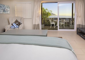 A bedroom with a bed, a chair, a table, and a view of the sea through a sliding glass door.