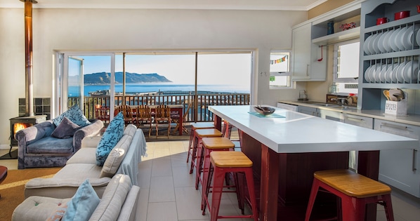 A modern kitchen with a large island, a dining area with red stools, and a view of the sea.
