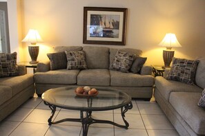 A living room with a sofa set, two side tables with lamps, a glass coffee table with a fruit bowl, and a framed picture on the wall.