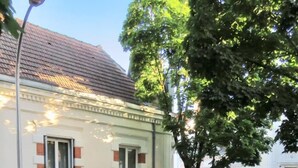 En bygning med hvid facade og røde vinduesrammer, en gade med biler og et træ i forgrunden.