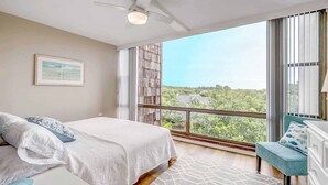 Un dormitorio con cama, ventilador de techo, ventana grande con vistas y una pequeña zona de estar.