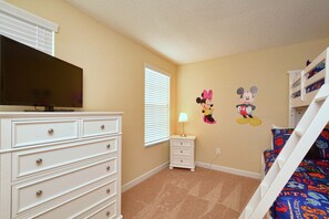 Habitación infantil con televisor, cómoda, mesita de noche, lámpara y un adhesivo de Mickey Mouse en la pared.