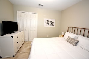 A bedroom with a bed, a dresser, a television, a nightstand, a lamp, and a door. - Greater Groves - 5 Bedrooms House w/pool-5202GG Home by RedAwning (Clermont)