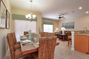 Un coin repas avec une table dressée pour quatre personnes, entouré de chaises en osier, d’un lustre et d’un ventilateur de plafond.