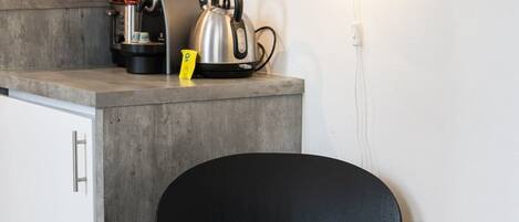A modern kitchen with a countertop, kettle, and kettle holder. There is a chair with a cushion and a wall-mounted light fixture.