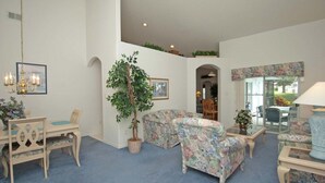 Ett vardagsrum med ett blått mattgolv, soffor med blommönster, ett matbord med stolar, en kristallkrona och ett stort träd i en kruka.