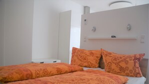 A hotel room with two beds, each with orange patterned bedding. There is a white wardrobe and a small shelf above the beds. - Nice apartment on the ground floor for 2 persons (Höxter (Lüchtringen))
