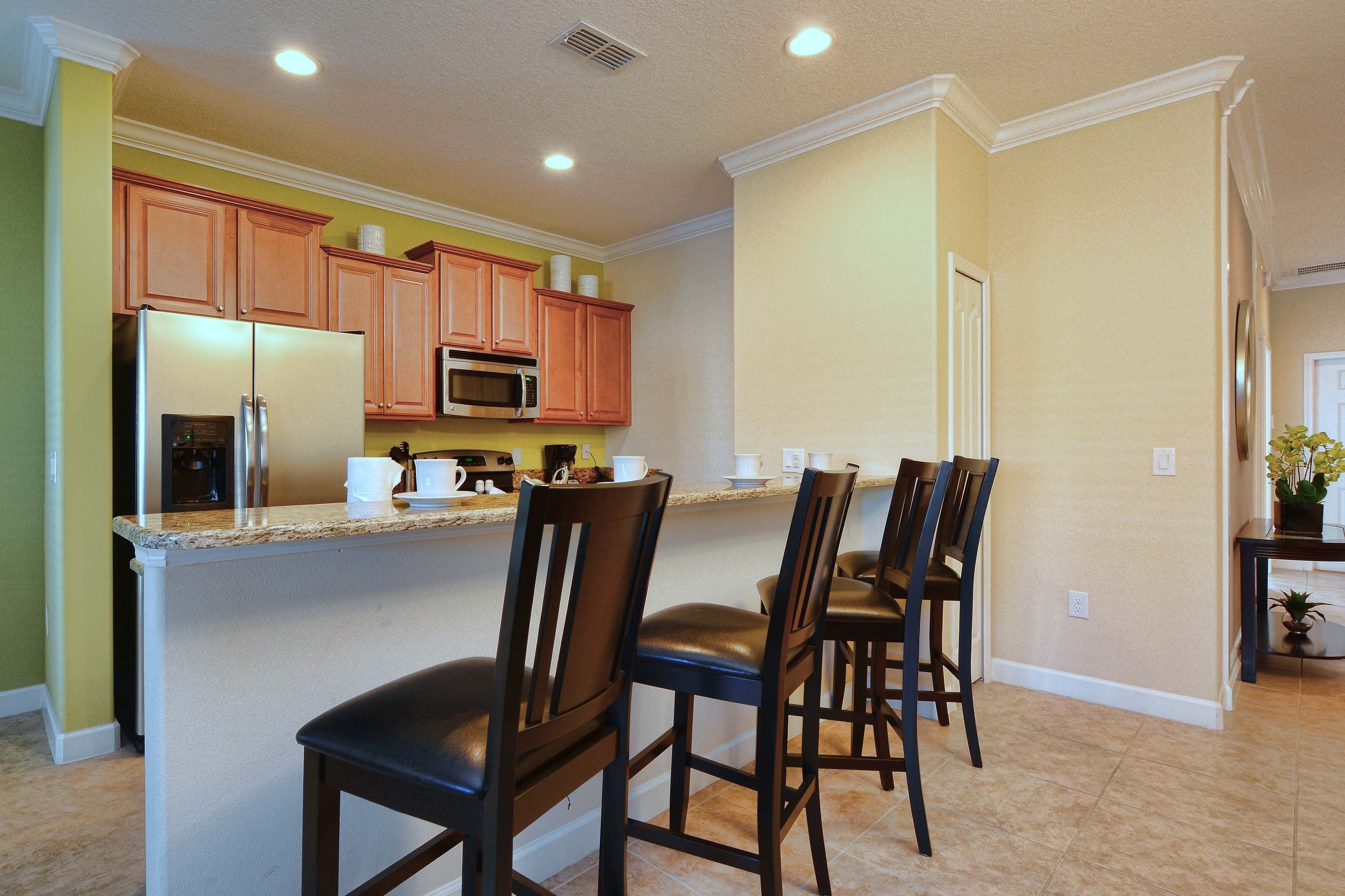 A kitchen with a built-in microwave, a refrigerator, and wooden cabinets. There are three bar stools and a small table with a potted plant.