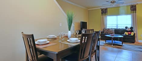 Un comedor con una mesa preparada para cuatro, sillas, un jarrón con planta y un televisor al fondo.