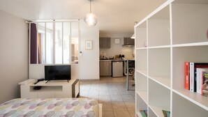 A compact living space with a bed, a TV, a bookshelf, and a kitchenette in the background. - Le cocon des Monts d'Or - cosy studio (Saint-Germain-au-Mont-d’Or)