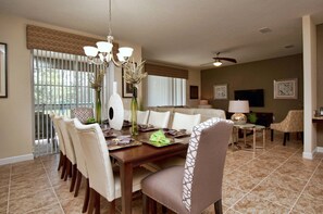 A dining area with a wooden table, chairs, and a chandelier. There is a sofa and a television in the background.