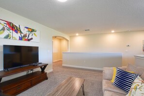A living room with a flat-screen TV on a wooden stand, a beige sofa with striped pillows, and a colorful wall art piece above the TV. - Solterra - 5 Bedrooms House w/pool-7035SR Home by RedAwning (Davenport)
