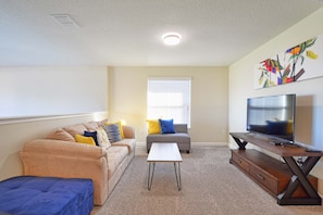 A living room with a beige sofa, a gray sofa, a blue ottoman, a coffee table, a TV on a wooden stand, and a window with blinds. - Solterra - 5 Bedrooms House w/pool-7035SR Home by RedAwning (Davenport)
