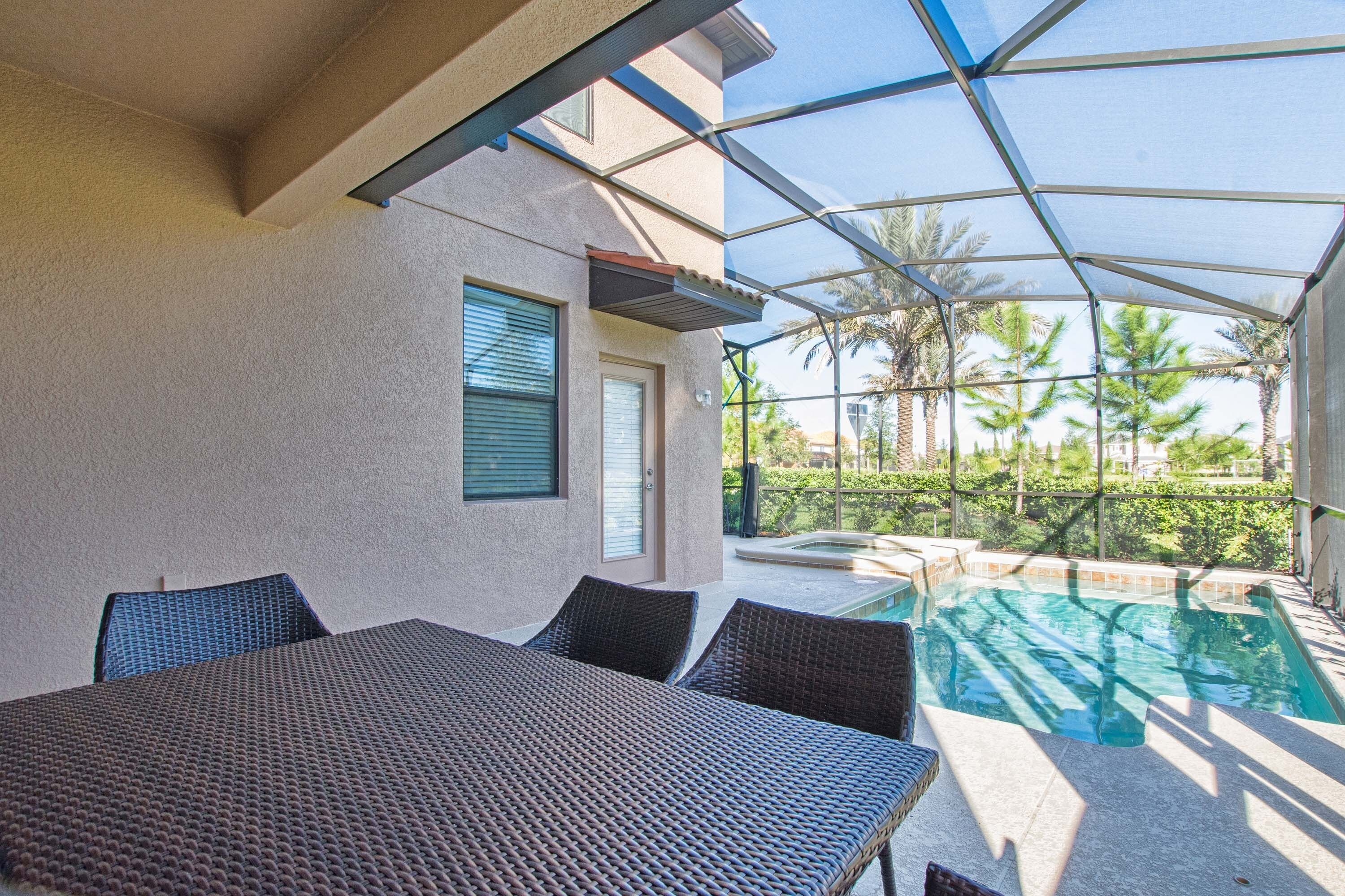 A covered outdoor area with a pool, a table with chairs, and a view of palm trees.