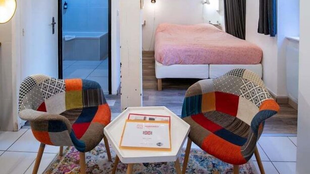 Ein modernes Zimmer mit einem Bett, zwei bunten Stühlen, einem kleinen Tisch und einer Deckenleuchte.