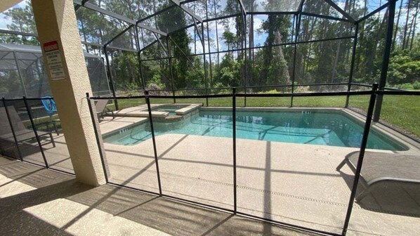 Un área de piscina con cerramiento de vidrio, solárium con baldosas y vista a árboles.