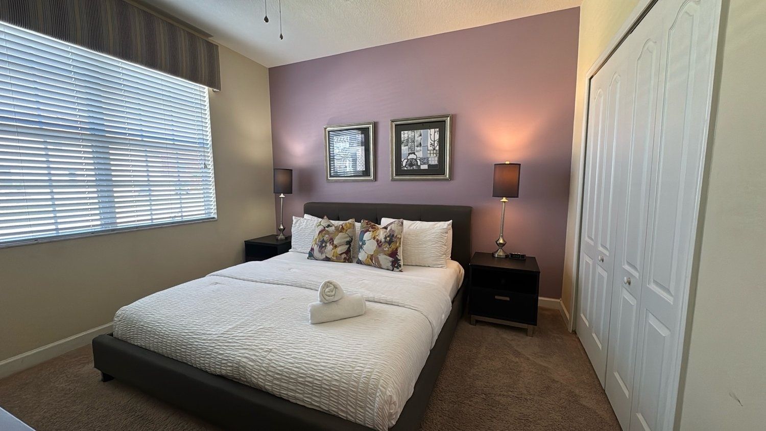 A bedroom with a bed, two bedside tables, a nightstand, a window with blinds, and a door.