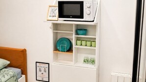 Quarto compacto com uma cama, uma prateleira branca fixada na parede com um micro-ondas e quadros decorativos.