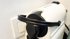 Um eletrodoméstico branco com painel de controle e pedal preto, provavelmente uma máquina de café, em frente a uma parede branca.