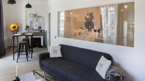 A modern living room with a dark sofa, two stools, a dining table with a lamp, and a large window with artwork. - Beautiful atypical apartment AC / garage (Lyon)