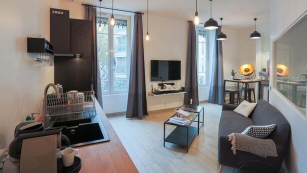 A modern living room with a sofa, coffee table, and a TV. There is a kitchen area with a coffee machine and cabinets. The room has large windows with curtains. - Beautiful atypical apartment AC / garage (Lyon)