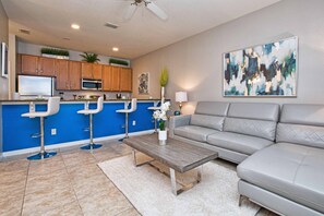 Un salon moderne avec un canapé gris, une table basse en bois et un coin cuisine aux accents bleus et aux meubles de rangement en bois.