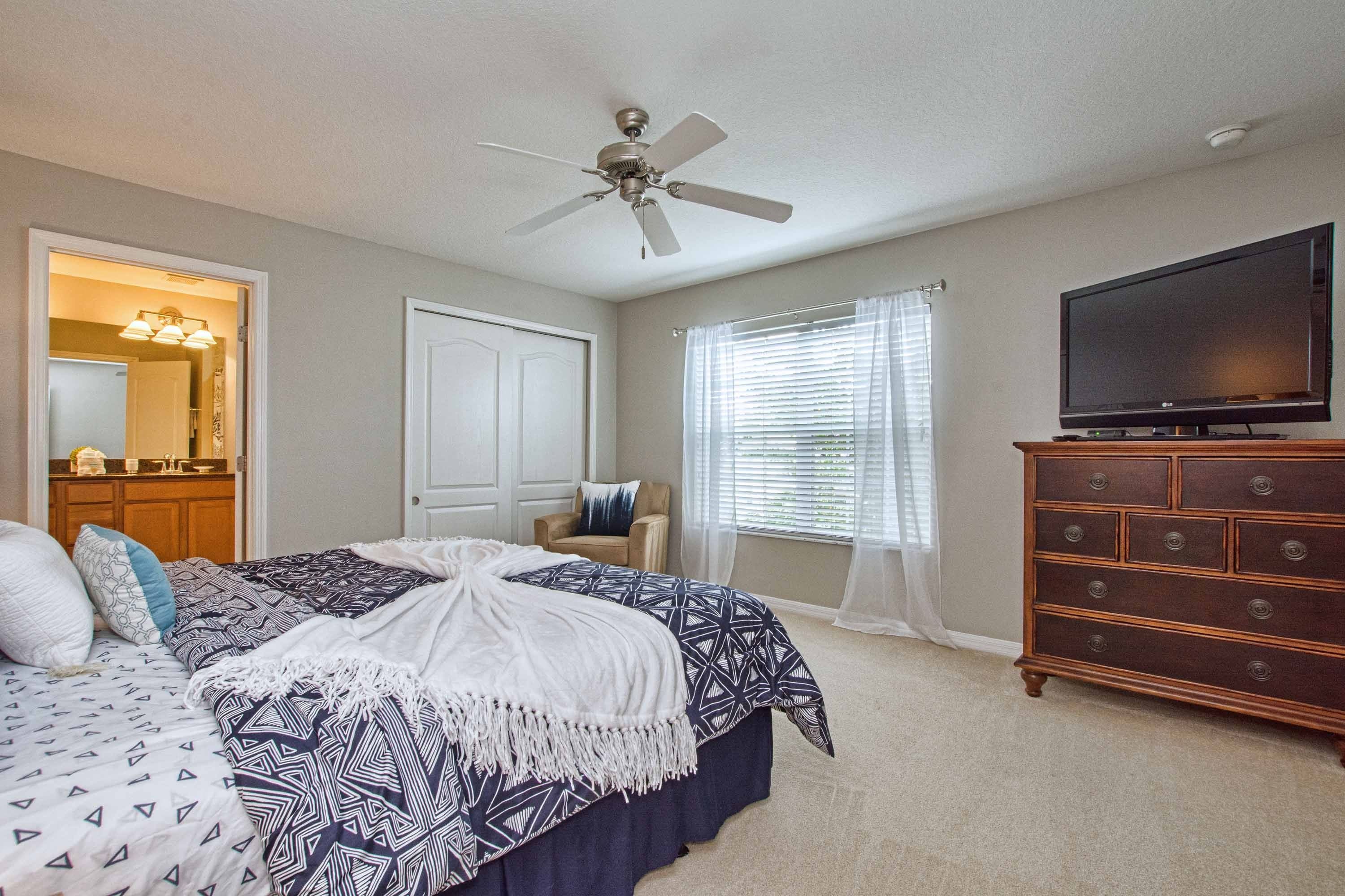 A bedroom with a bed, a dresser, a television, a ceiling fan, and a window with blinds.