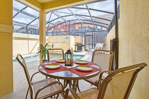 Eine überdachte Terrasse mit Tisch für zwei Personen, mit Blick auf einen Swimmingpool und ein Gebäude mit rotem Dach.