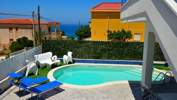 A swimming pool with lounge chairs, a view of the sea, and a yellow building in the background. - Brand new Villa With Pool and Alcamo Marina Terrace (Alcamo)