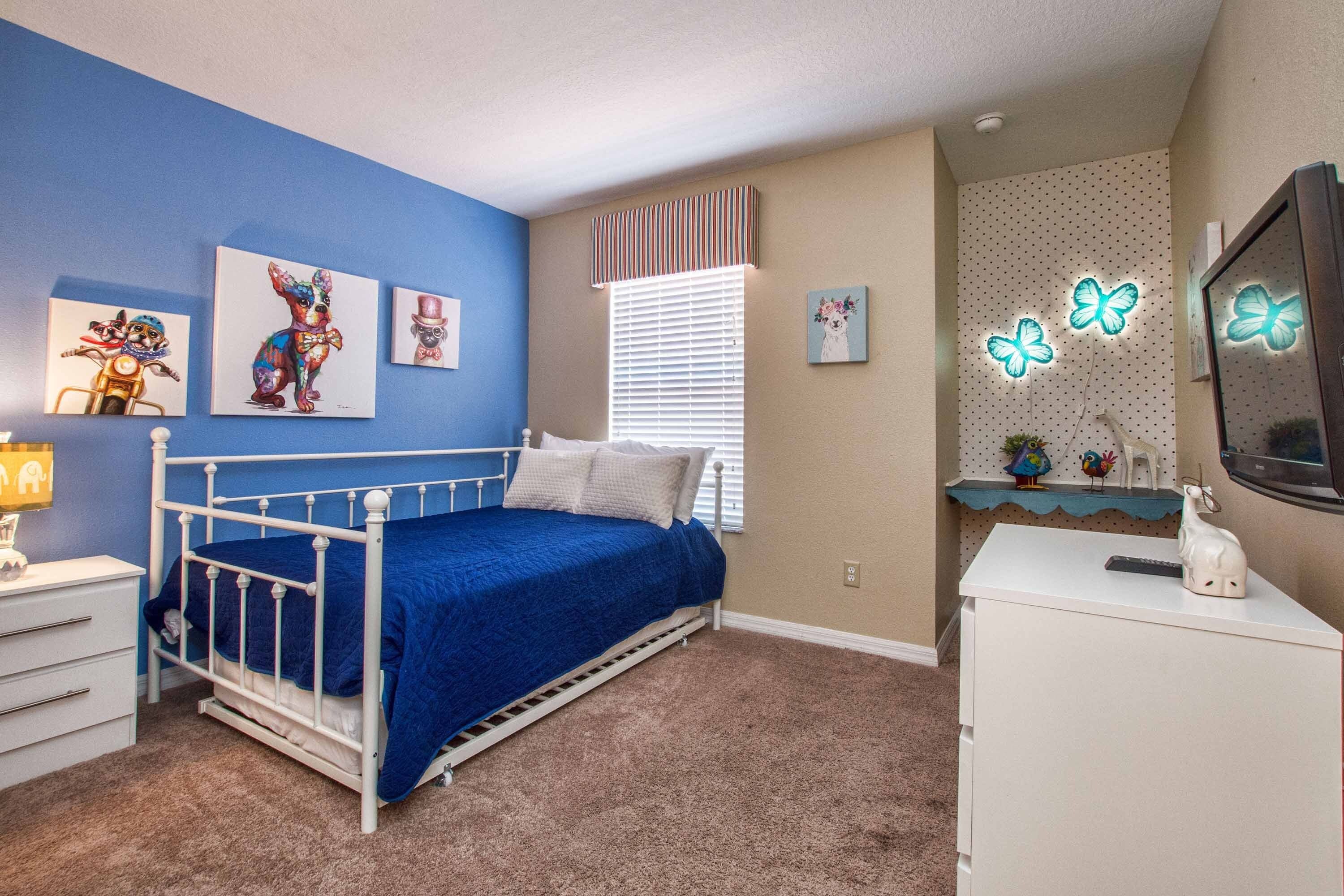 Quarto com cama de metal branca, roupa de cama azul, cômoda branca, escrivaninha com computador e decoração de parede, incluindo pinturas de cachorros e luminárias em formato de borboleta.