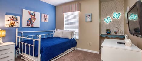 Quarto com cama de metal branca, roupa de cama azul, cômoda branca, escrivaninha com computador e decoração de parede, incluindo pinturas de cachorros e luminárias em formato de borboleta.
