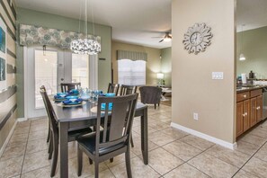 A dining area with a table set for four, a kitchen with wooden cabinets, and a living area with a couch and a lamp. - Paradise Palms- 4 Bed Townhome W/splashpool-3057pp 4 Bedroom Townhouse by RedAwning (Kissimmee)